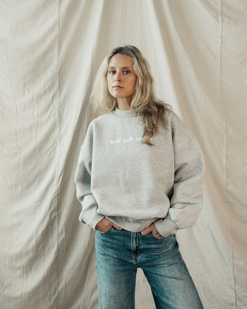 Woman wearing a light grey oversized sweatshirt from the Soft Top Co. Comfort Collection, standing in front of a soft draped fabric backdrop.