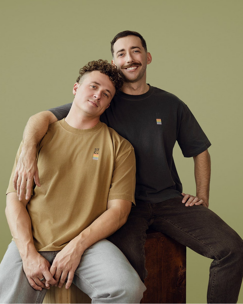 Two people wearing Soft Top Co. Proud & Soft Collection tees sit together against a muted green backdrop. One leans comfortably on the other’s shoulder, both relaxed and smiling, showcasing the soft, minimal design of the shirts.